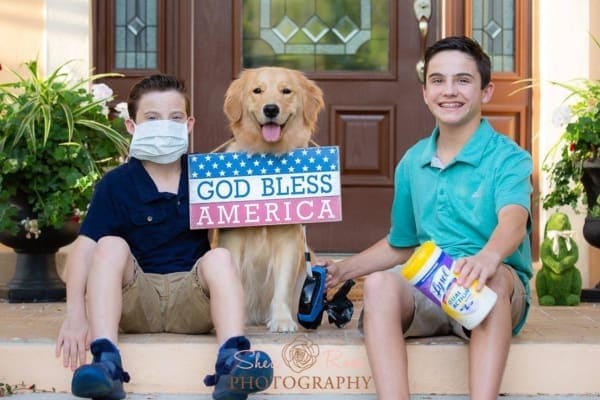 two kids sitting on front porch with dog, container of lysol, face mask, sign saying god bless america. photo. 